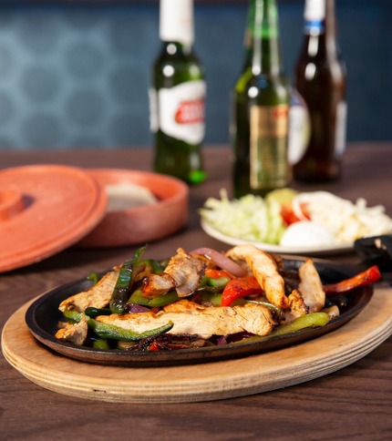 Sizzling platter of fajitas with grilled chicken strips, bell peppers, and onions, accompanied by beer and tortillas in Casa Amigos