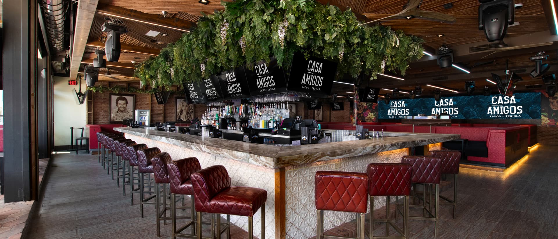 central wooden bar countertop, red quilted bar stools, hanging greenery, and flat-screen TVs